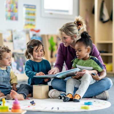 A group of children in a daycare or classroom setting engages with a teacher reading a book to them. The playroom background fosters an atmosphere of learning and playful interaction.