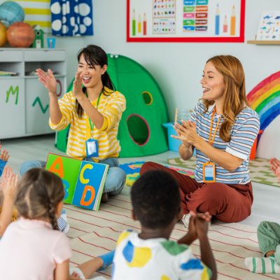 A preschool teacher sits on the floor of her classroom with a small group of students as she reads them a book.