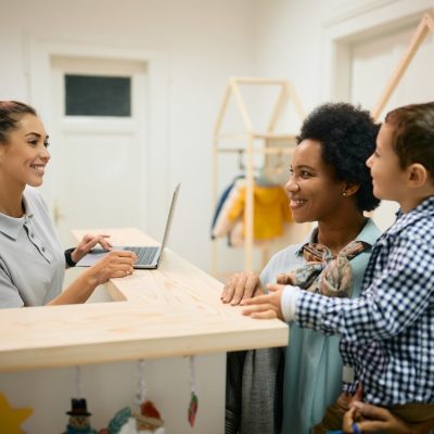 Happy kindergarten teacher communicating with African American mother and her son at reception desk.